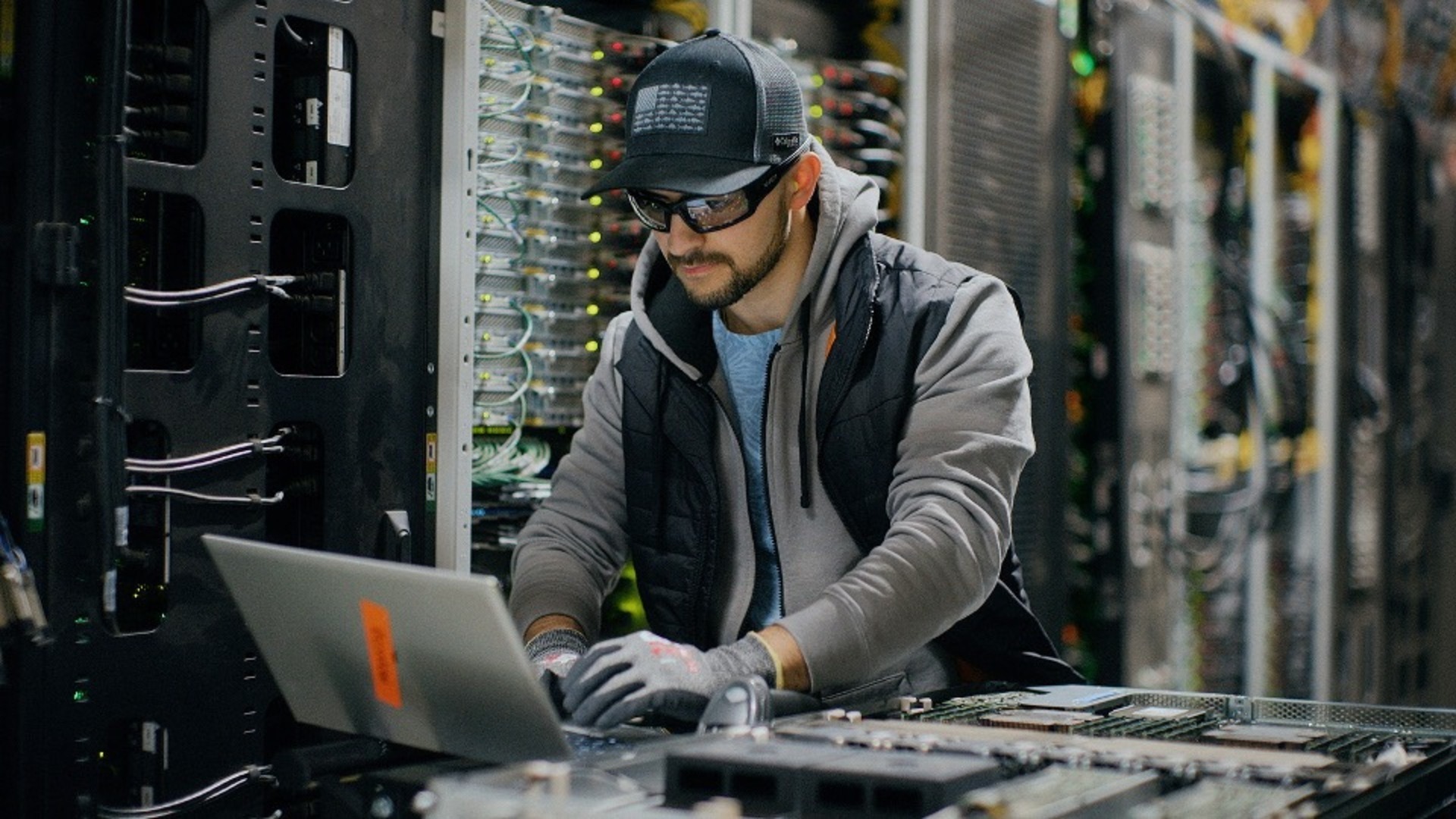 Technician working on a laptop in a server room surrounded by equipment racks.